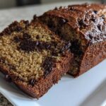 A freshly baked loaf of chocolate chip banana bread, partially sliced, showing moist interior texture and melted chocolate chips.