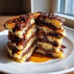 A stack of fluffy chocolate chip pancakes cut in half, showing layers of melted chocolate and syrup.