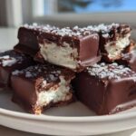 Stack of chocolate-covered cool whip candy squares topped with flaky sea salt, one piece is bitten.