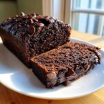 A loaf of rich, dark Chocolate Zucchini Banana Bread with a slice cut, topped generously with melted chocolate chips.