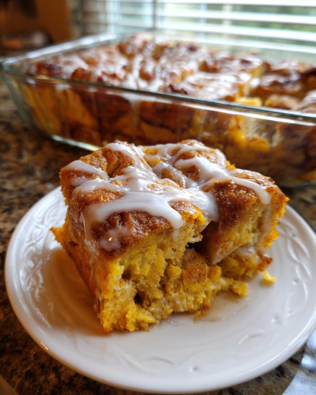 A square serving of cinnamon roll french toast bake topped with white icing on a white plate.