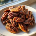 A pile of glistening Cinnamon Sugar Roasted Pecans coated in sugar crystals, served on a white plate near a window.