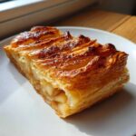 A close-up of a golden, flaky slice of classic apple puff pastry on a white plate.