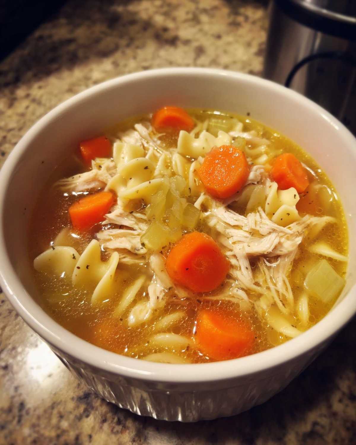 Close-up of a white bowl filled with rich broth, shredded chicken, egg noodles, and bright orange carrots, perfect chicken noodle soup.