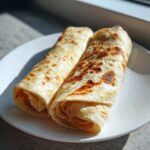 Two golden-brown, rolled cottage cheese wrap tortillas sitting on a white plate in bright sunlight.