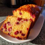A slice of moist Cranberry Orange Loaf Bread showing bright red cranberries embedded in the yellow crumb, next to the rest of the loaf.