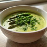 A close-up of a bowl of creamy asparagus soup, topped with whole asparagus spears and fresh chopped parsley.