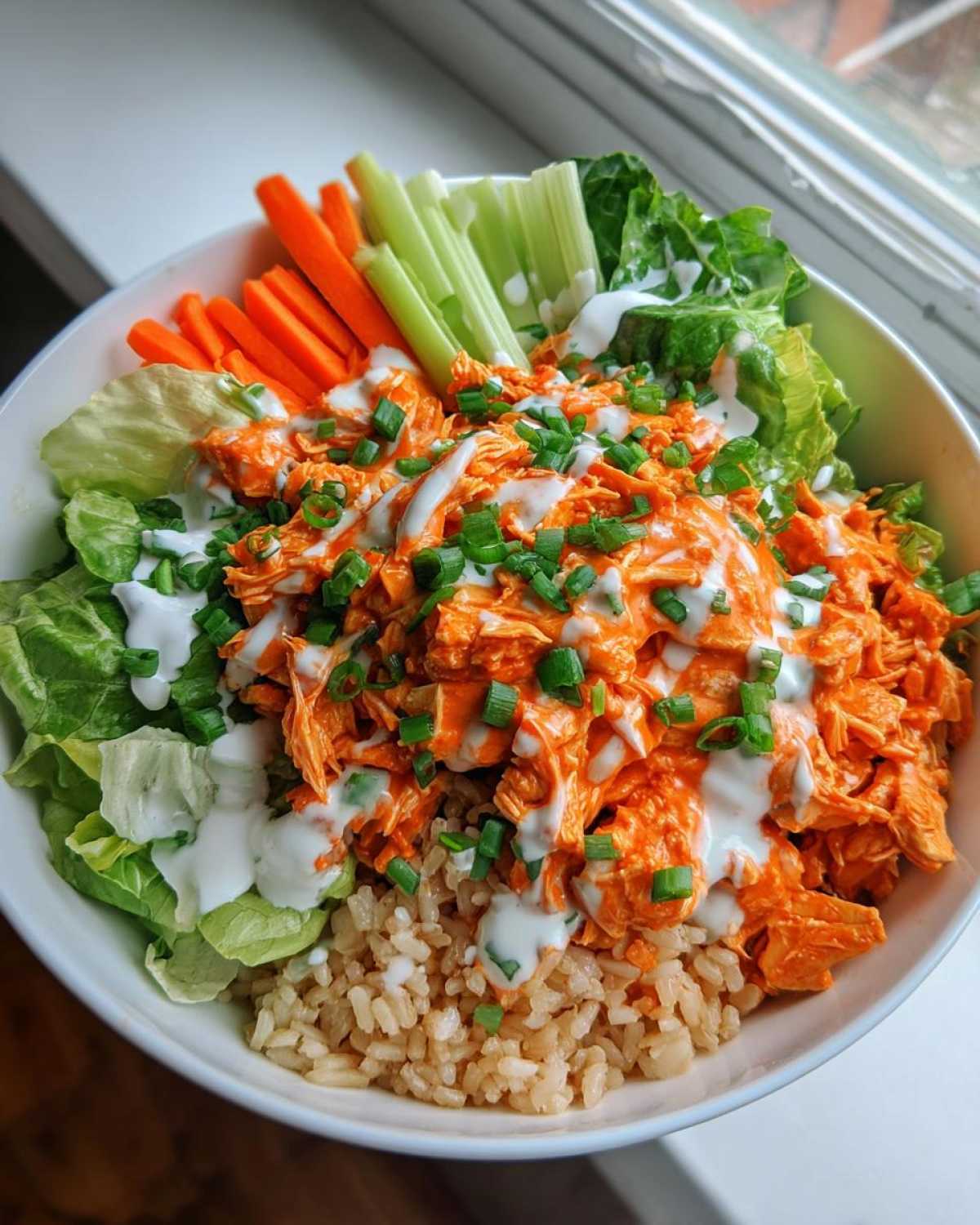 A vibrant buffalo chicken bowl featuring shredded buffalo chicken, brown rice, lettuce, carrots, celery, and a drizzle of ranch dressing.