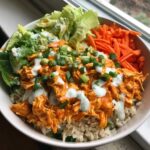 A close-up of a creamy buffalo chicken bowl featuring shredded chicken, brown rice, lettuce, carrots, and ranch dressing.