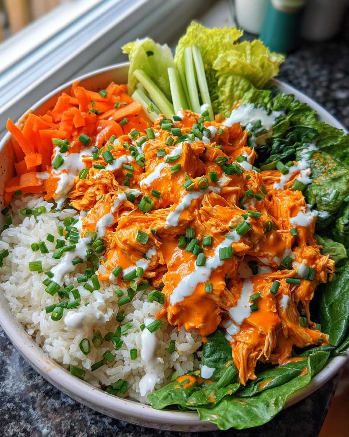 A vibrant buffalo chicken bowl featuring shredded buffalo chicken, white rice, carrots, celery, lettuce, and a ranch drizzle.