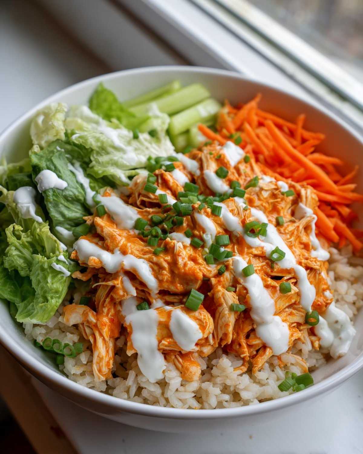 Close-up of a creamy buffalo chicken bowl featuring shredded buffalo chicken over brown rice, topped with ranch dressing, carrots, celery, and scallions.