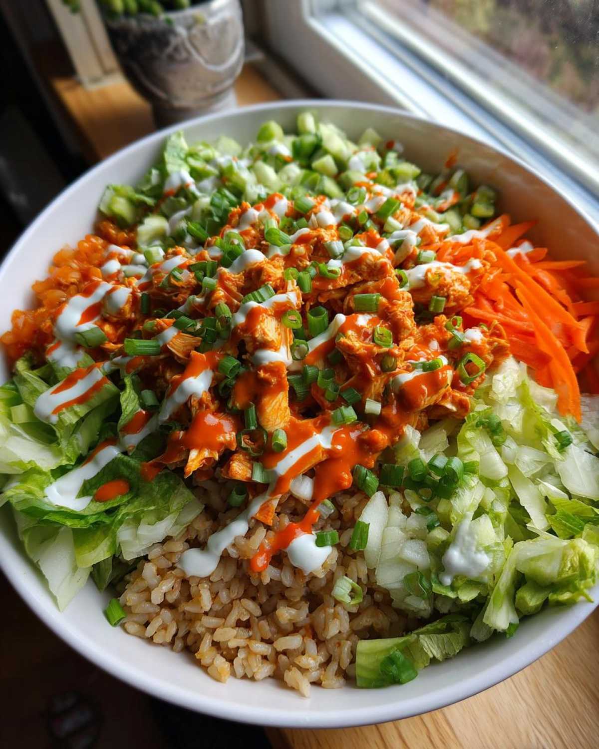 A vibrant buffalo chicken bowl featuring shredded chicken tossed in sauce, brown rice, lettuce, carrots, and drizzled with ranch dressing.