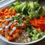A vibrant buffalo chicken bowl featuring shredded chicken tossed in orange sauce, drizzled with ranch, over brown rice, surrounded by lettuce, carrots, and cucumbers.
