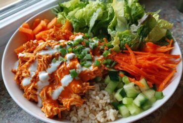 A vibrant buffalo chicken bowl featuring shredded chicken tossed in orange sauce, drizzled with ranch, over brown rice, surrounded by lettuce, carrots, and cucumbers.