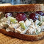 Close-up of a creamy chicken salad sandwich piled high on whole wheat bread with red grapes and celery.