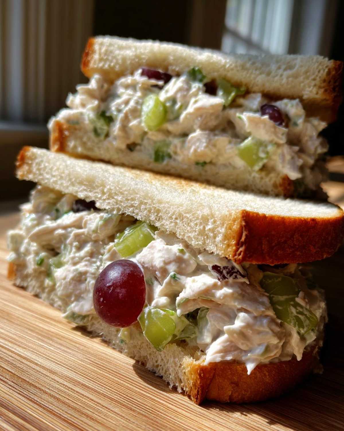 Close-up of a stacked chicken salad sandwich featuring creamy filling, green celery, and red grapes on white bread.