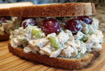 Close-up of a creamy chicken salad sandwich piled high on whole wheat bread with red grapes and celery.