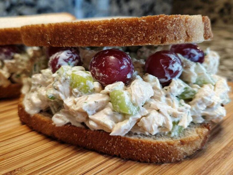 Close-up of a creamy chicken salad sandwich piled high on whole wheat bread with red grapes and celery.