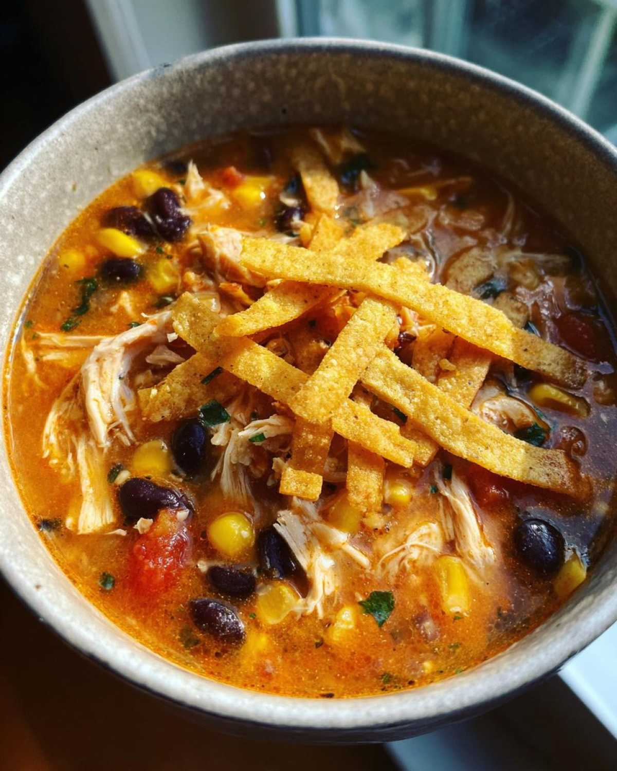 Close-up of a bowl of creamy chicken tortilla soup, featuring shredded chicken, black beans, corn, and crispy tortilla strips.