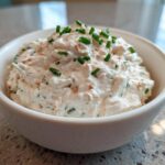 A close-up of creamy clam dip served in a white bowl, garnished generously with chopped green chives.
