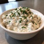 A close-up of a white bowl filled with creamy clam dip, generously topped with chopped green chives.