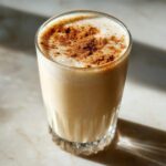 A tall glass of creamy, light brown coquito topped with a generous dusting of ground cinnamon.