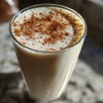 A tall glass filled with creamy coquito, topped with a thick layer of foam and dusted heavily with ground cinnamon.