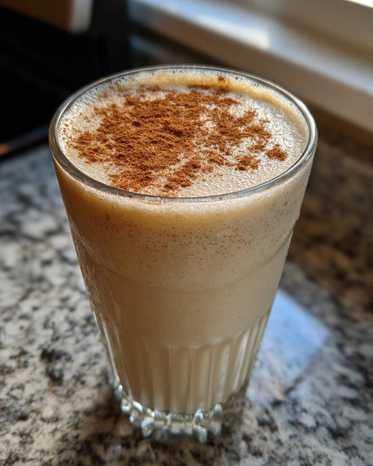 A close-up of a glass filled with creamy, frothy coquito, generously dusted with ground cinnamon on top.