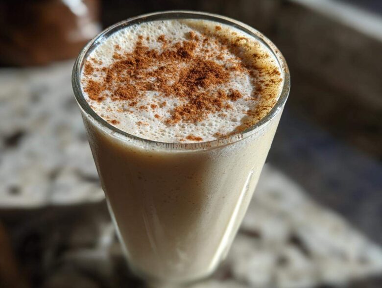 A tall glass filled with creamy coquito, topped with a thick layer of foam and dusted heavily with ground cinnamon.