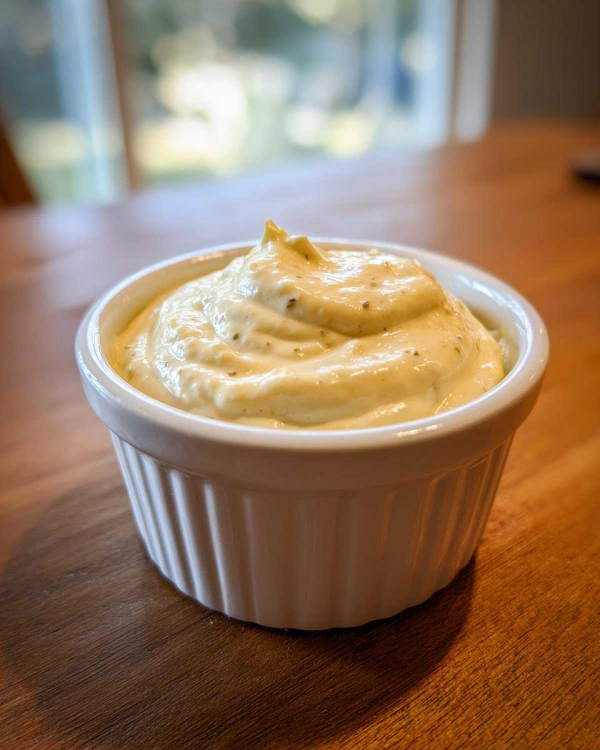 A close-up of thick, creamy garlic aioli, speckled with herbs, served in a small white fluted ramekin on a wooden table.