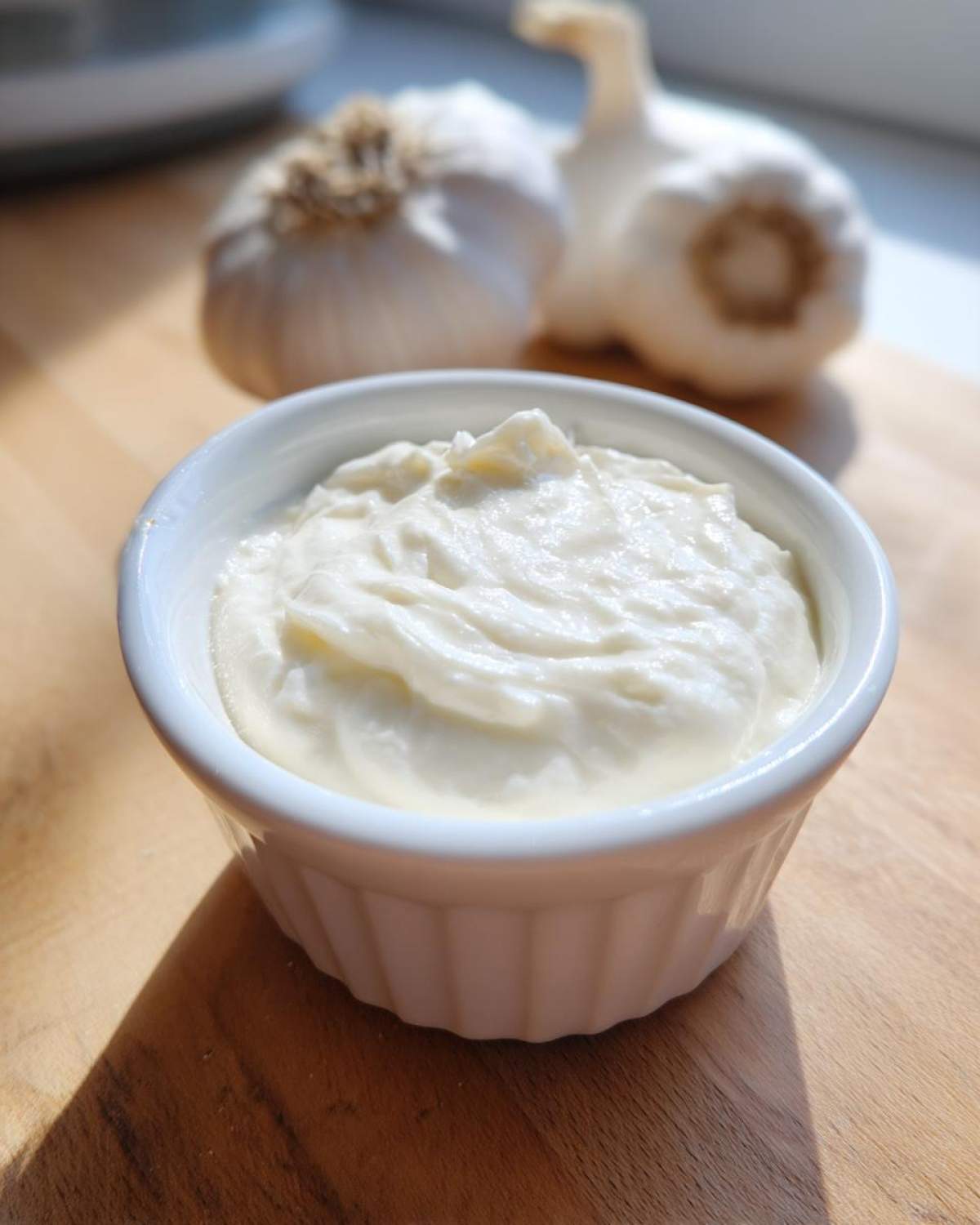 A small white ramekin filled with thick, creamy garlic sauce, with two whole garlic bulbs blurred in the background.