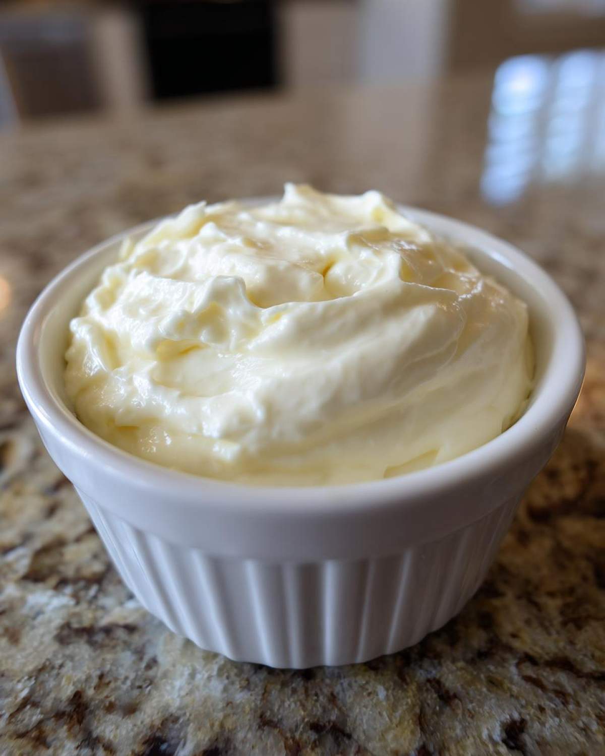 A close-up of thick, creamy garlic sauce served in a small white fluted ramekin on a granite countertop.