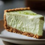 A close-up of a thick slice of creamy, light green key lime cheesecake on a white plate, topped with lime zest.