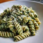 Close-up of fusilli protein pasta coated in a creamy green spinach or pesto sauce, topped with grated cheese.