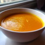 Close-up of a white bowl filled with rich, orange Creamy Pumpkin Soup resting on a windowsill.