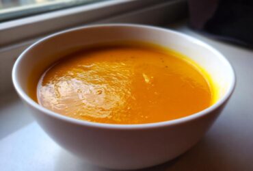 Close-up of a white bowl filled with rich, orange Creamy Pumpkin Soup resting on a windowsill.