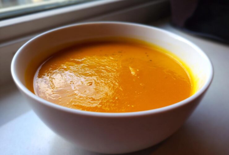 Close-up of a white bowl filled with rich, orange Creamy Pumpkin Soup resting on a windowsill.