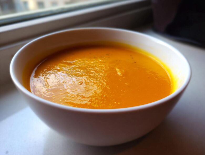 Close-up of a white bowl filled with rich, orange Creamy Pumpkin Soup resting on a windowsill.