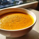 Close-up of a white bowl filled with vibrant orange Creamy Pumpkin Soup, topped with freshly cracked black pepper.