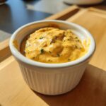 A close-up of thick, orange-hued remoulade sauce speckled with green herbs, served in a small white ramekin.