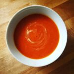 Overhead view of a white bowl filled with vibrant orange-red Tomato Soup, featuring a swirl of cream on top, set on a wooden table.