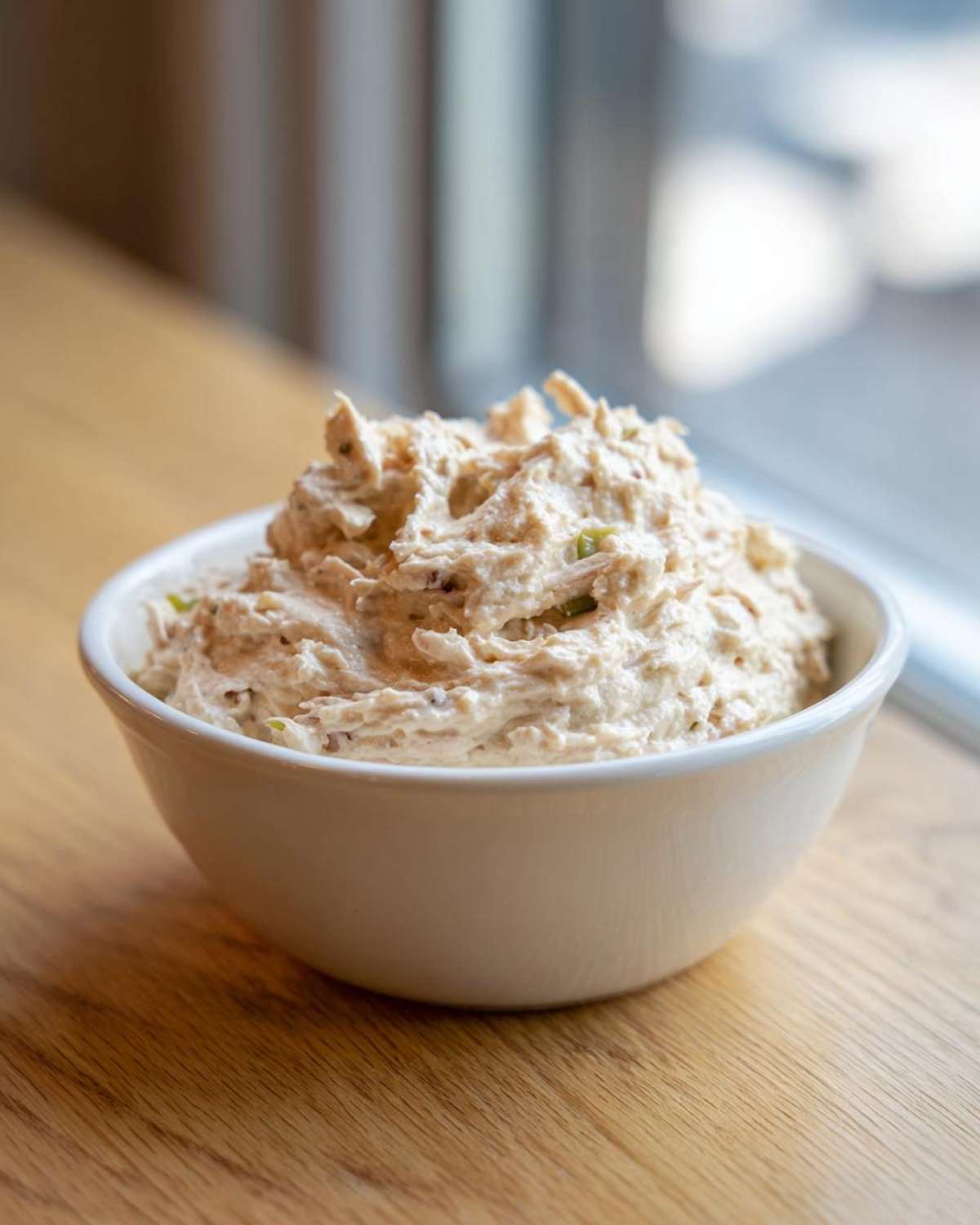 A white bowl filled with creamy, textured tuna dip, set on a wooden surface near a window.
