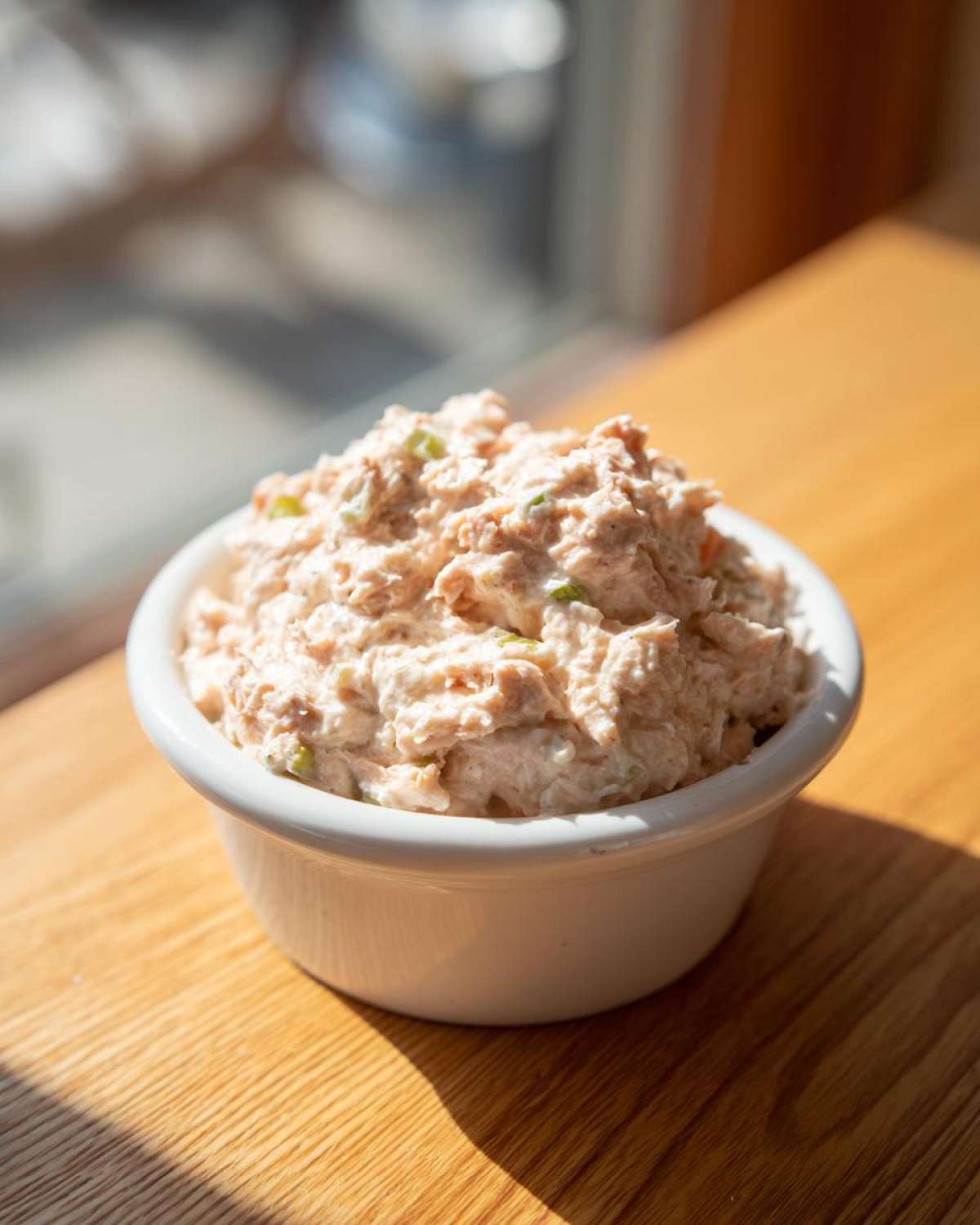A small white bowl filled with creamy, pinkish tuna dip, sitting on a sunlit wooden table.