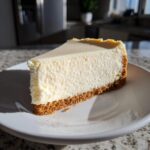 A perfect slice of creamy white chocolate cheesecake with a graham cracker crust sitting on a white plate.