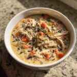 A close-up of a creamy bowl of amazing wild rice soup, featuring shredded chicken, carrots, celery, and a thyme garnish.
