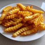 A close-up of golden brown, crispy crinkle cut air fryer french fries piled high on a white plate.