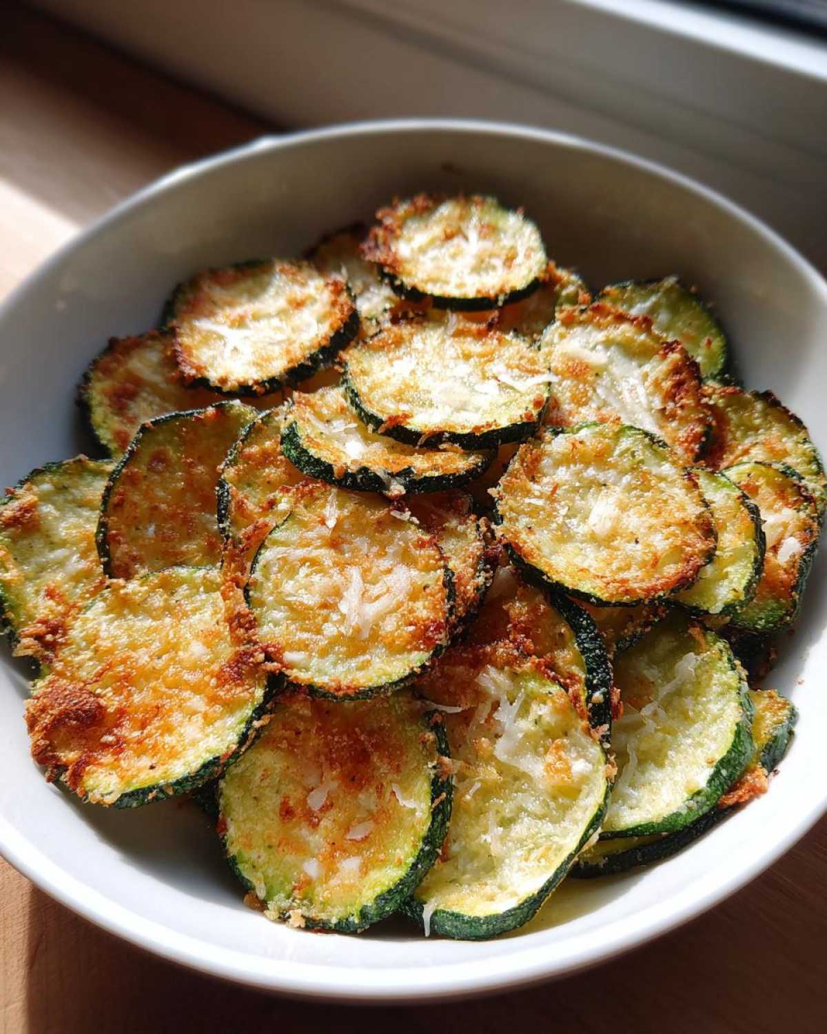A white bowl filled with golden brown, crispy zucchini chips topped with Parmesan cheese.