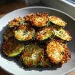 A close-up of a plate piled high with golden brown, crispy zucchini chips coated in Parmesan cheese.