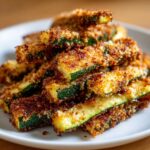 A close-up of a pile of golden-brown, crispy zucchini fries coated in breadcrumbs on a white plate.