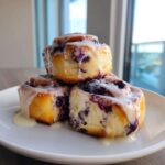 Three decadent blueberry cheesecake rolls stacked on a white plate, drizzled generously with white icing.